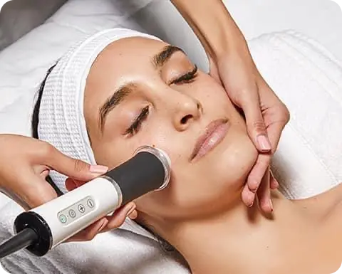 Ženska med neinvazivnim tretmajem obraza, kjer strokovnjak uporablja aparat na predelu ličnic, medtem ko z drugo roko podpira brado.