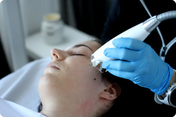 Ženska med neinvazivnim tretmajem obraza, kjer strokovnjak z rokavicami uporablja aparat na predelu ličnic.